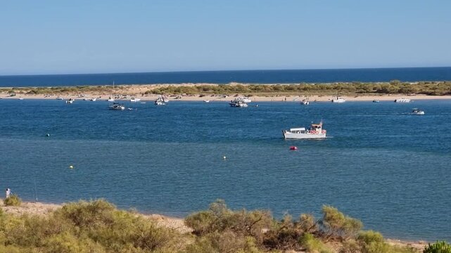 Wide clip of a small ferry and boats cruising the Piedra River mouth near Flecha del Rompido sandbar in Cartaya, Huelva, Spain, under a clear blue sky.