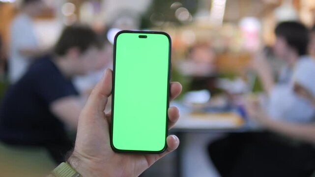 Hand holds smartphone with green screen in cafe. Background shows blurred people dining. Footage ideal for app previews or digital marketing. Focus stays on phone for clear mockup use