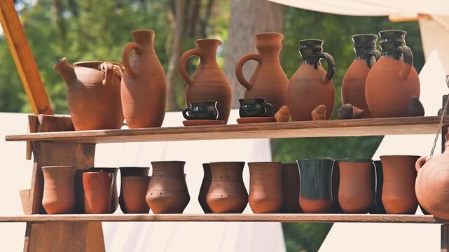 Craftsmen work on pottery, shaping clay into jugs and bowls at a historical reenactment. A variety of earthenware is displayed on wooden shelves in a natural outdoor setting