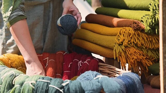 A person sorts various colored yarn from a woven basket in a medieval marketplace. The setting features textiles of different hues arranged neatly, with a focus on craft and trade
