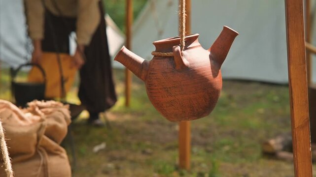 In a medieval camp, a wooden kettle hangs from a post while a person dressed in simple tunic and pants tends to bags nearby. The background shows tents and tools used for daily tasks