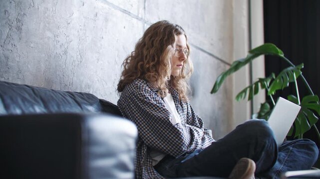 Focused Caucasian woman using laptop while sitting on sofa in modern home office. Curly haired female working remotely and concentrating on online project. Comfortable workspace indoors.