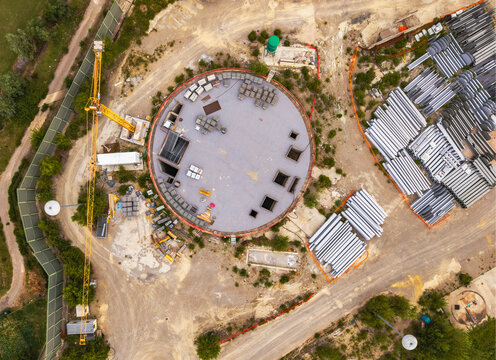 Aerial view of a large circular construction site featuring a tall yellow crane, organized stacks of metal pipes, and various building materials in Serravalle Scrivia, Piedmont, Italy.