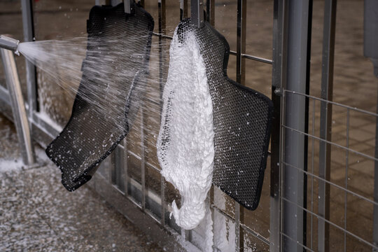 High pressure water stream spraying foam off black EVA car floor mats hanging on metal holders at self service car wash.