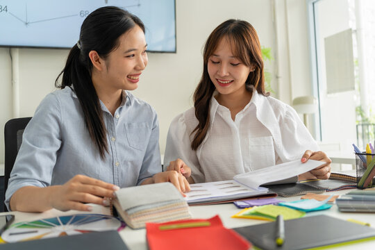 Two Asian women working together in a bright, modern studio.while using a laptop and smartphone. The creative workspace is filled with colorful samples for seasonal palette selection.