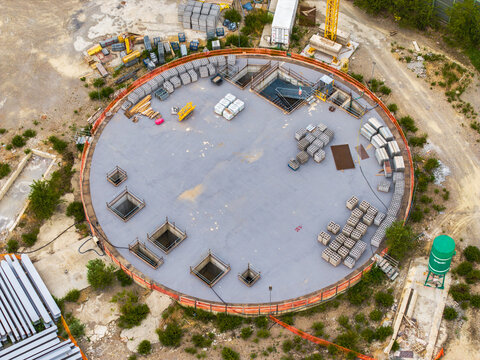 Aerial view of a circular construction site with stacks of bricks, metal frames, and a green silo on the ground in Serravalle Scrivia, Piedmont, Italy.