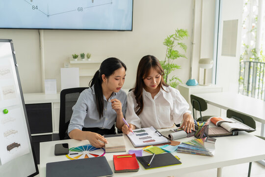 Two Asian women working together in a bright, modern studio.while using a laptop and smartphone. The creative workspace is filled with colorful samples for seasonal palette selection.