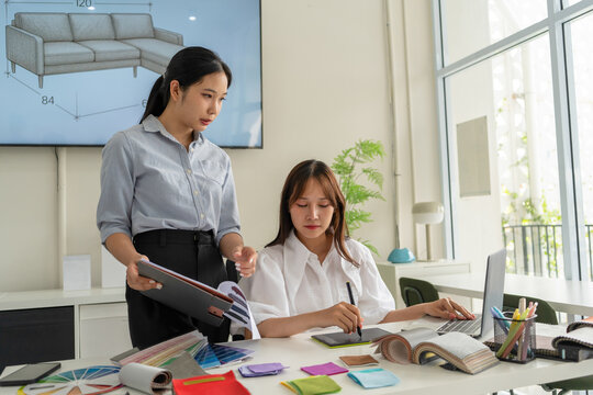 Two Asian women working together in a bright, modern studio.while using a laptop and smartphone. The creative workspace is filled with colorful samples for seasonal palette selection.