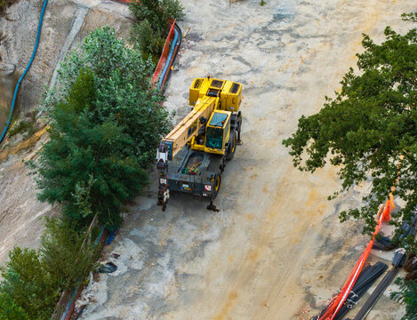 Aerial view of a yellow mobile crane parked on a construction site with orange safety fencing, construction materials, and surrounding trees in Serravalle Scrivia, Piedmont, Italy.