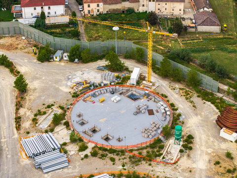 Aerial view of a circular construction site featuring a yellow tower crane, stacks of building materials, and concrete foundations, Serravalle Scrivia, Piedmont, Italy.