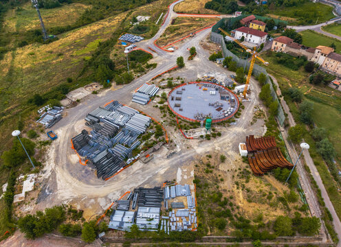 Aerial view of a construction site featuring stacks of industrial materials, a circular foundation, and a yellow crane in Serravalle Scrivia, Piedmont, Italy.