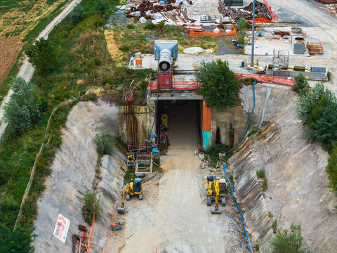 Aerial view of the construction site at the tunnel entrance with yellow excavators, industrial equipment, and surrounding vegetation in Serravalle Scrivia, Piedmont, Italy.