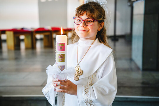 Child girl in traditional white first communion clothing holding a candle inside a Catholic church during a religious celebration.