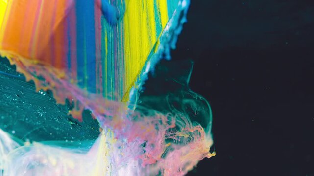 Close-up paint slab edge shedding multicolored pigment cloud with glossy teal surface and dark negative background