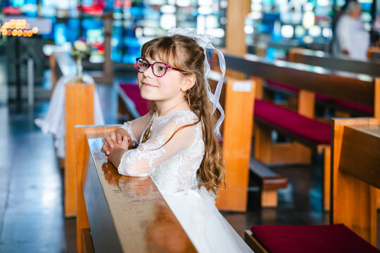 Child girl in traditional white first communion dress kneeling in prayer with folded hands inside a Catholic church during a religious celebration.