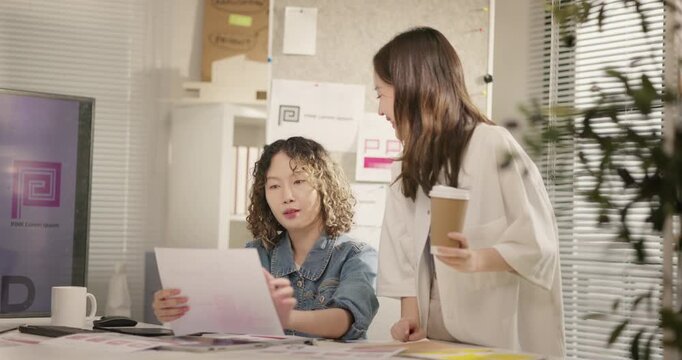 Asian female graphic designers brainstorm design ideas at studio desk. Two business woman working together on branding project over a visual layout. creative marketing, teamwork, coordination concept.
