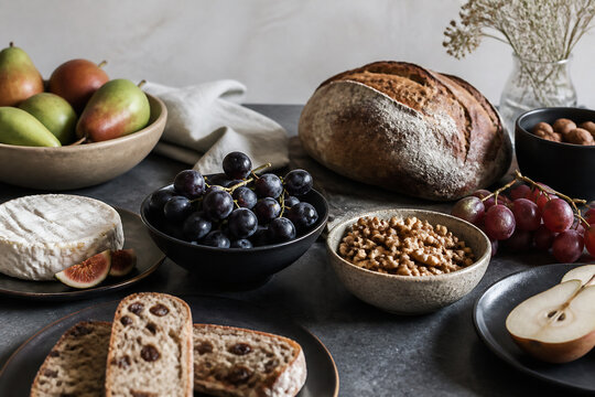 A rustic still life arrangement featuring bread, fruit, cheese, nuts, and grapes on a dark surface.