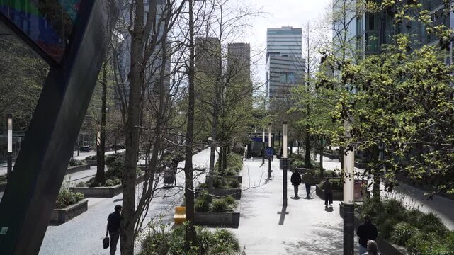A high-angle view of a tree-lined pedestrian boulevard in Amsterdam's Zuidas business district, featuring modern office skyscrapers, walkways, and commuters strolling on a sunny day.