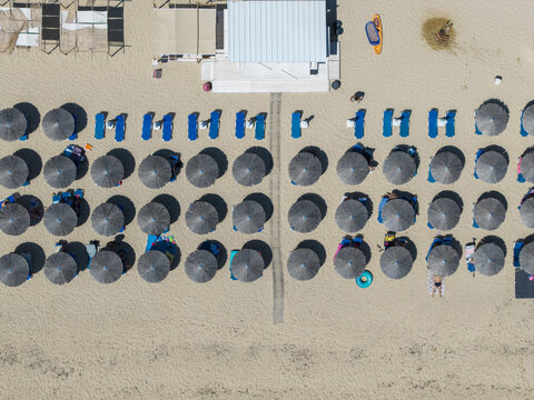 Aerial view of Kriaritsi Beach featuring rows of straw umbrellas and blue sunbeds on the sandy shore with people relaxing in Greece.