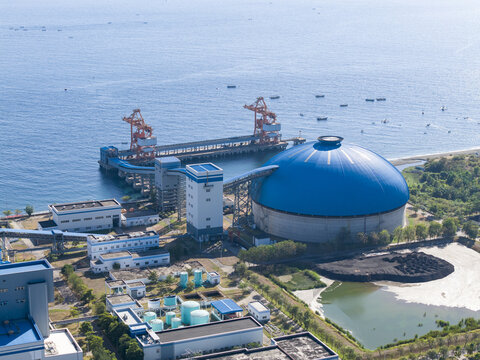 Aerial view of Celukan Bawang Steam Power Plant featuring a large blue dome, industrial infrastructure, and coal storage facilities along the coastline in Buleleng, Bali, Indonesia.