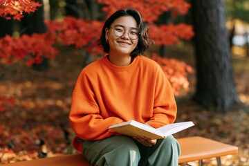 Fototapeta premium Smiling student reading book on park bench in autumn foliage for seasonal background and banner design