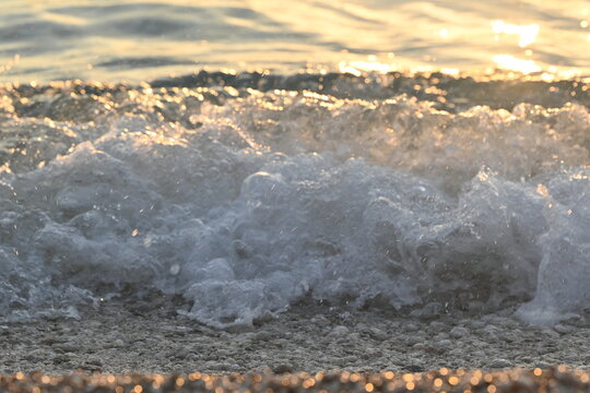  Sea wave with white foam crashing on pebble beach under golden sunset light close up background