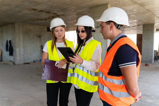 Builder and inspectors consult on floor plan of project. Female specialists and construction worker look at tablet studying dimensions of structures