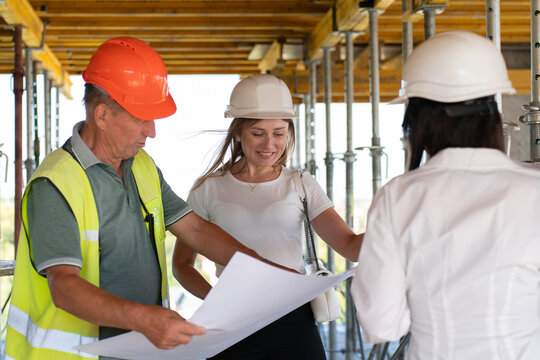 Construction engineers review blueprints working together at building site. Inspectors and foreman discuss plan at unfinished structure