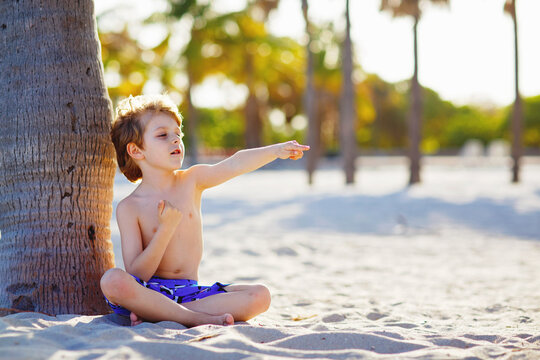 Blond adorable gorgeous little kid boy having fun on Miami beach, Key Biscayne. Happy healthy cute child playing with sand and running near ocean. Palms, security house and white sand