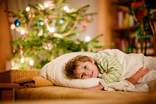 Little cute blond boy sleeping under Christmas tree and dreaming of Santa at home, indoors. Traditional Christian festival. Happy kid child waiting for gifts on xmas. Cozy soft light