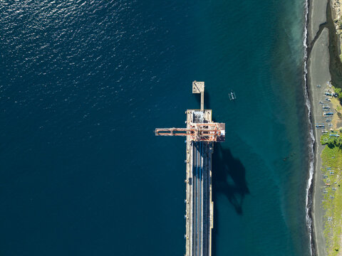 Aerial view of Celukan Bawang Steam Power Plant with its industrial pier extending into the deep blue sea near the dark sand coastline in Buleleng, Bali, Indonesia.