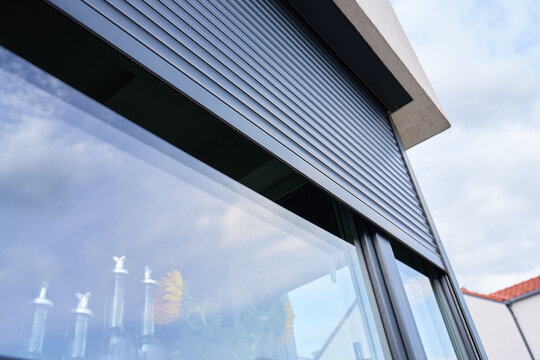 Roller shutter partly covers modern house window, with clouds reflected in the glass