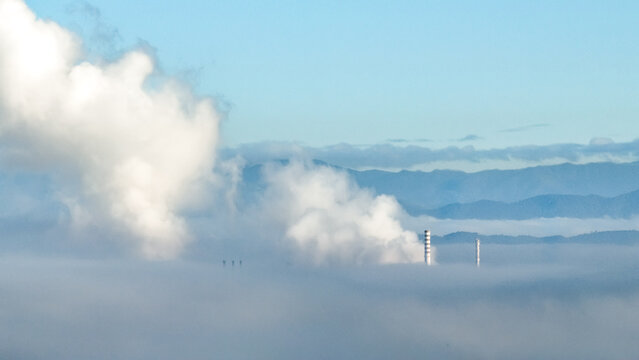 Aerial view of industrial chimneys emitting thick smoke above a layer of low-lying clouds with mountains in the background in Brazil.