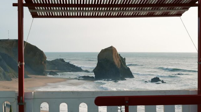 Horizontal 4K video showing the classic red canopy bench on a coastal promenade with Portuguese pavement, looking out at dramatic sea stacks and ocean waves in Santa Cruz, Portugal