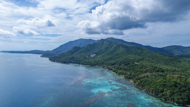 Aerial view of a lush green island surrounded by clear turquoise water and coral reefs with mountains in the background under a blue sky with clouds in Indonesia.