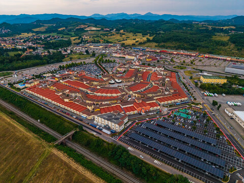 Aerial view of Designer Outlet Serravalle with its distinctive red rooftops, extensive parking areas with solar panels, and surrounding green hills in Serravalle Scrivia, Piemonte, Italy.