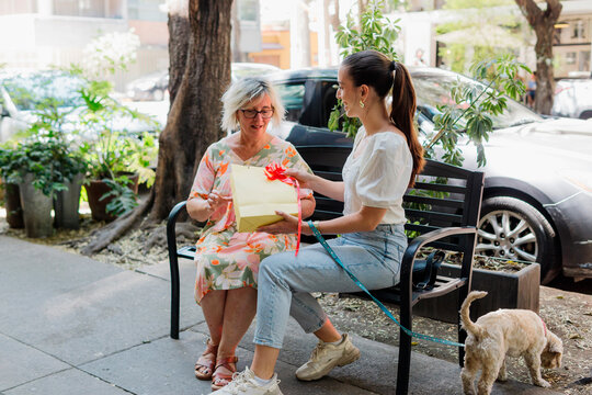 A woman that giving a gift to her mother on a street bench.