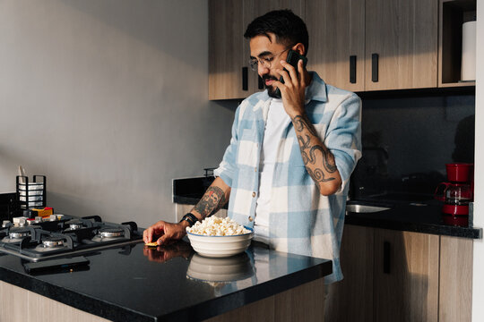 Young man talking on his cell phone and eating snacks in the kitchen