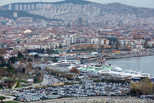 Aerial view of the coastal harbor featuring numerous ferries docked at the pier, a large parking lot, and dense urban buildings stretching towards the hills in the background, Istanbul, Turkey.