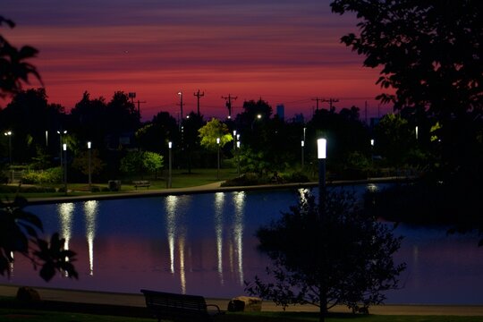 Twilight purple and red sky reflected in Scissortail Park pond with glowing lamp posts at dusk in Oklahoma City