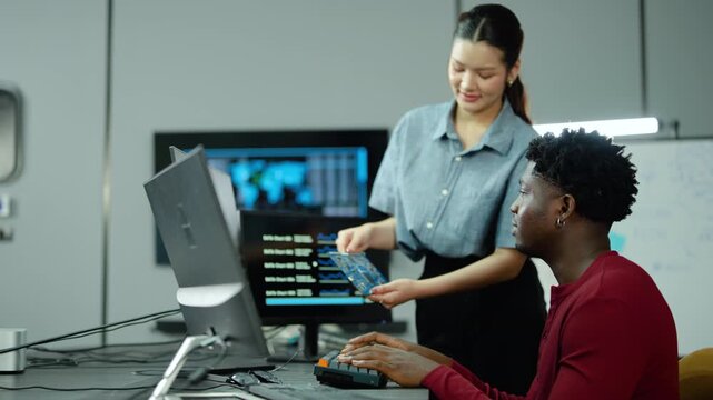 Asian female and Black male engineers collaborate in tech laboratory, analyzing microchip circuit board and computer monitor data during hardware research, development, and motherboard innovation.