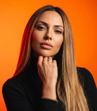 Gorgeous woman in black turtleneck posing with hand under chin on bright orange background.