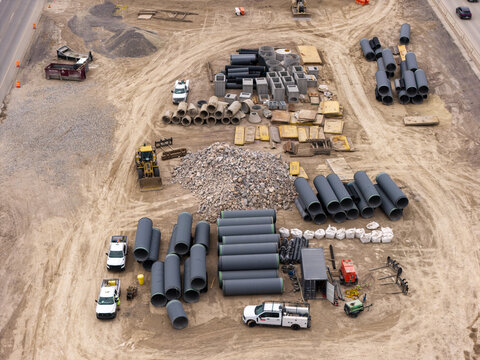 Aerial view of the 2100 North Freeway construction project featuring stacks of large plastic and steel pipes, construction vehicles, and gravel piles on a dirt site in Lehi, Utah, United States.