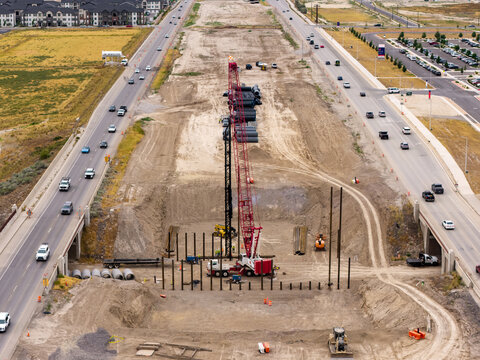 Aerial view of the 2100 North Freeway construction project featuring a large crawler crane, deep excavation, and steel foundation piles near suburban roads in Lehi, Utah, United States.
