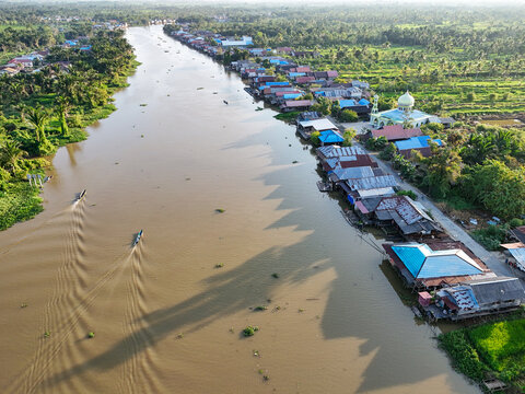 Aerial view of the riverfront village of Sungai Tabuk with traditional houses, a mosque, and boats navigating the waterway in Sungai Tabuk, South Kalimantan, Indonesia.