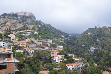 Fototapeta premium Terracotta rooftops of Castelmola village clustered on mountainside seen through soft morning fog in Sicily. Concept of layered village architecture and atmospheric hilltop community photography.
