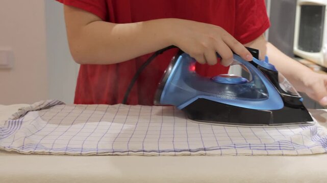 A person smooths a checked dish towel with a corded iron on a padded ironing board in a home kitchen.