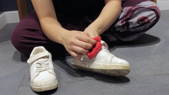 Hands remove dirt from a white sneaker using a red sponge while another shoe lies nearby.