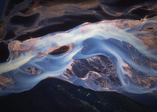 Aerial view of a braided glacial river with vibrant blue water flowing through dark volcanic sand and sediment in Iceland.