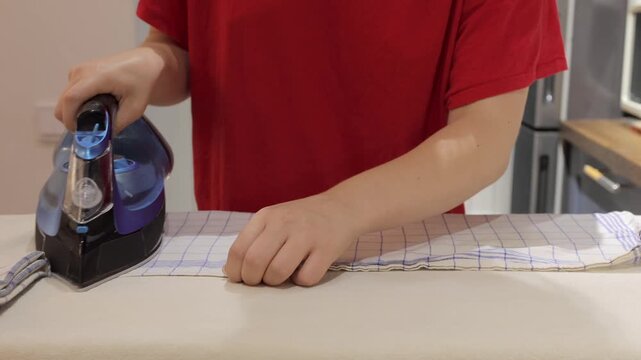 A checked cloth is pressed with a corded iron as hands adjust the edge on an ironing board.